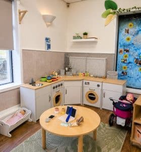 The home corner in Bright Blossoms - play kitchen, dolls and a pram beside the round table